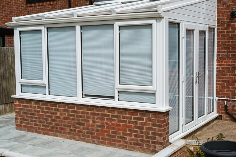 White lean to conservatory with blinds on all windows and doors, seen from the outside on a sunny day