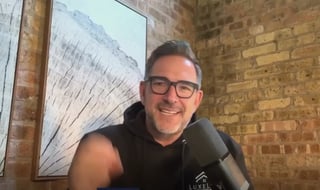 Man wearing glasses and black shirt at microphone in front of brick wall with framed artwork