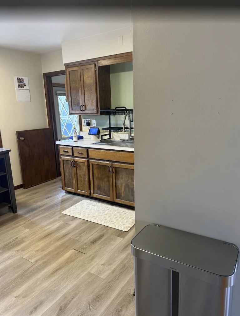 Kitchen area with wooden cabinets, stainless steel trash bin in foreground, and computer setup visible on counter through doorway