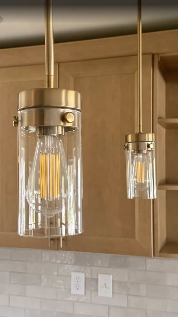 Two modern brass and glass cylinder pendant lights hanging over a white subway tile backsplash in a contemporary kitchen with tan cabinetry