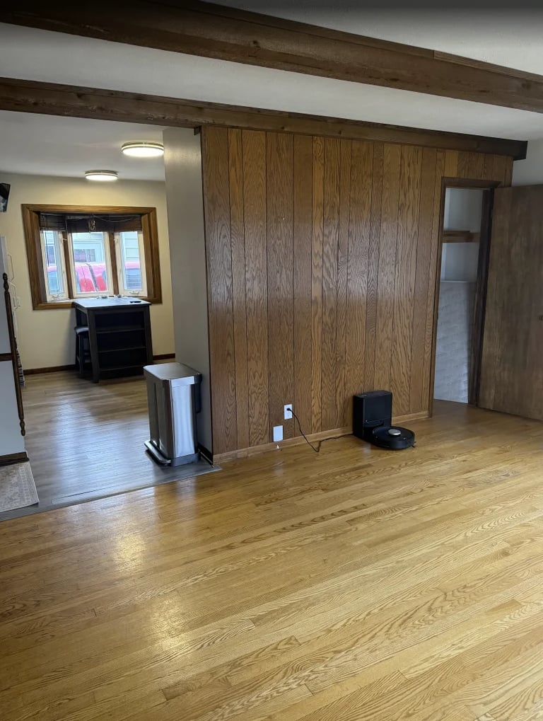 Empty living room with wood paneling, exposed beams, hardwood floors, and an open doorway leading to another room