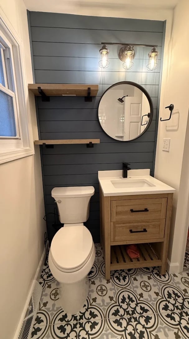 Modern bathroom with dark gray shiplap wall, wood vanity, round black mirror, white toilet, patterned tile floor, and industrial pendant lights