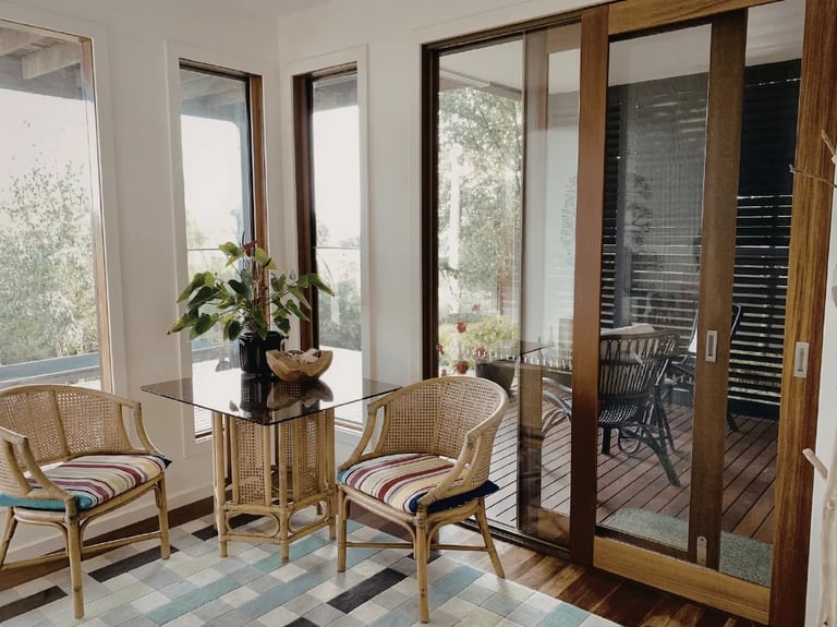 Modern sunroom with rattan armchairs, small round table with flowers, and glass doors opening to an outdoor deck with wooden flooring