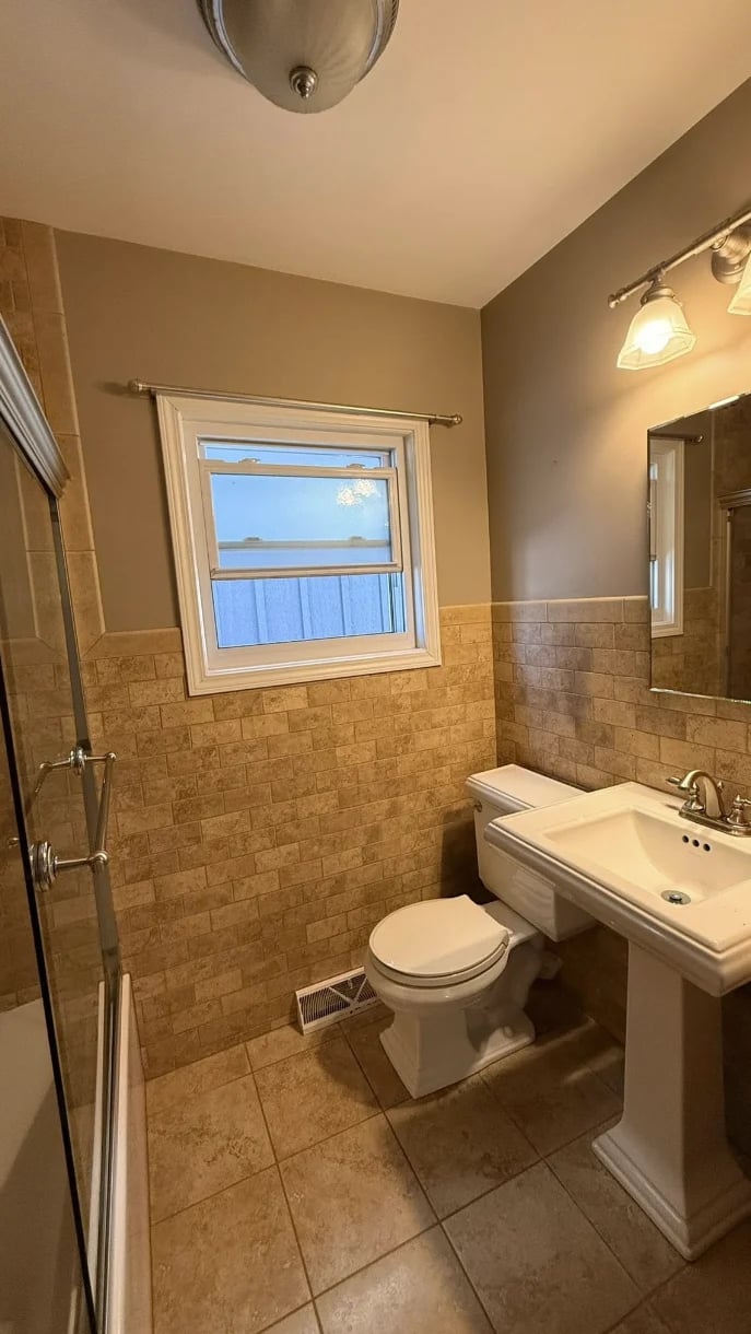 Bright bathroom with beige tile walls, white pedestal sink, toilet, and shower with glass enclosure. Window with blue sky view above toilet.