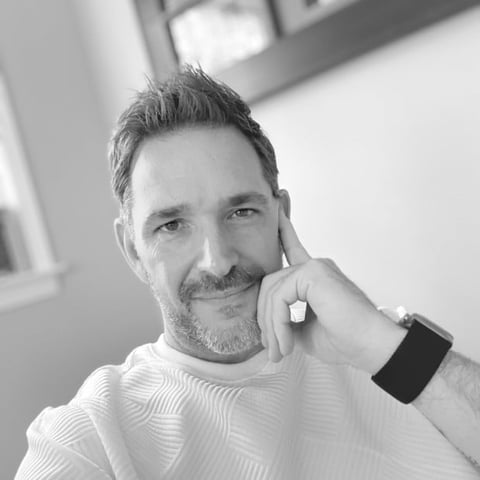 Black and white portrait of a man with dark hair and facial hair wearing a white textured sweater and smartwatch, tilting his head thoughtfully